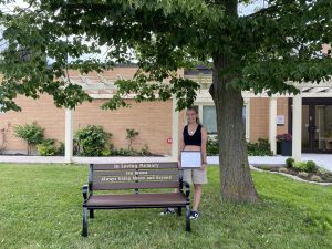 The scholarship award recipient stands next to the memorial bench that is placed outside beside a tree