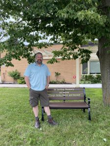 Ken Himbury is standing next to the Jen Brown memorial bench outside beside a tree